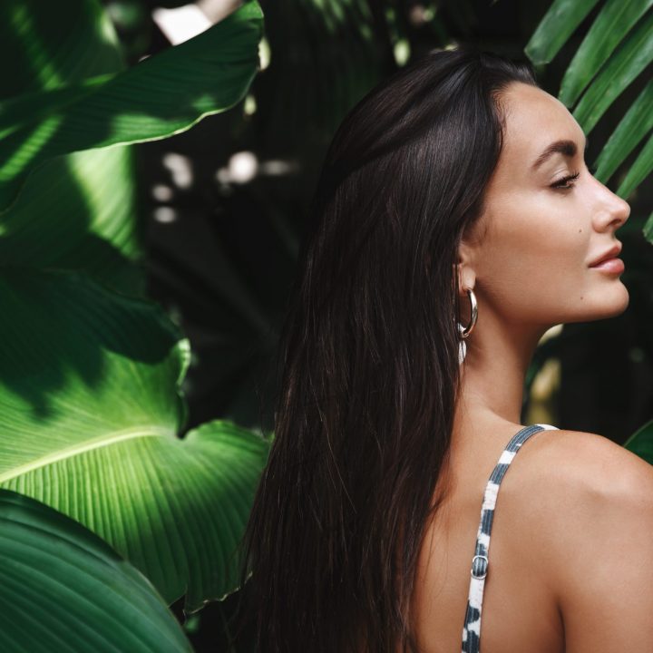 Tropical vacation and travelling concept. Rear view of beautiful brunette woman with golden tan, wearing bikini, standing in profile in green leaves, palm trees and tropic nature.